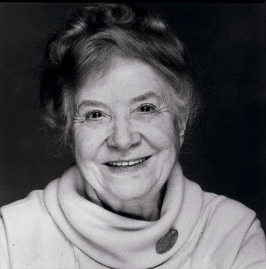 Retrato em preto e branco de Doris Allen, senhora idosa de cabelos claros presos para trás, sorrindo abertamente para a câmera, de blusa clara de gola alta com um broche oval.